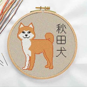 Akita Inu cross stitch pattern in a wooden embroidery hoop with colorful threads, featuring a standing Akita dog in orange and white next to the text Akita Inu in Japanese kanji