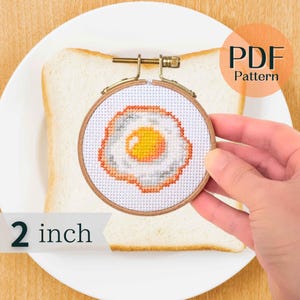 Puede incluir: Un aro de bordado de 5 cm de diámetro con un diseño de punto de cruz de un huevo frito sobre tela blanca. El aro está en un plato blanco con una rebanada de pan blanco.