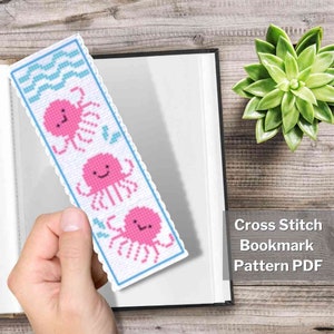May include: A cross stitch bookmark pattern featuring three pink octopuses with blue waves and a white background. The pattern is printed on a white paper with a blue border. The text "Cross Stitch Bookmark Pattern PDF" is printed in black at the bottom of the page.