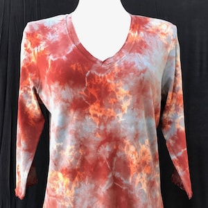 Op de afbeelding: Een tie-dye shirt met een V-hals en 3/4 mouwen. Het shirt is een mix van oranje, rood en blauwe kleuren.