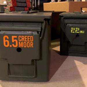May include: Two green metal ammo cans with labels. The can on the left has an orange label that says "6.5 CREEDMOOR". The can on the right has a yellow label that says "22 LR".