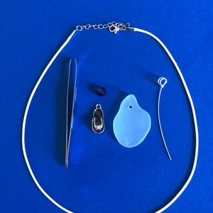 May include: A collection of jewelry-making supplies on a blue background. Includes a white cord necklace, a pair of tweezers, a small silver charm, a blue bead, a light blue sea glass pendant, and a wire with a loop.