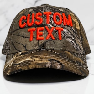 May include: A camouflage baseball cap with the words "CUSTOM TEXT" embroidered in bright orange. The hat features a curved brim and a classic six-panel design, perfect for outdoor activities.