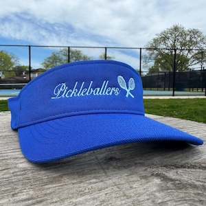 May include: A royal blue visor with the word "Pickleballers" in cursive script and two crossed pickleball paddles. The visor is sitting on a wooden surface, with a tennis court and trees in the background.