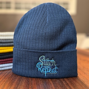May include: A navy blue knit beanie with a ribbed texture and the text "Stitch and Repeat" embroidered in green and blue.