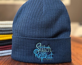 May include: A navy blue knit beanie with a ribbed texture and the text "Stitch and Repeat" embroidered in green and blue.