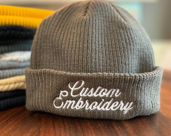 May include: A stack of gray, blue, beige, and yellow knit beanies with a gray beanie on top that has white embroidery that says "Custom Embroidery".