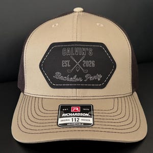 May include: A tan and brown trucker hat with a black patch that reads "CALVIN'S EST. 2026 Bachelor Party" with crossed golf clubs. The hat has a curved brim and an adjustable strap.