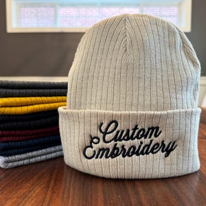 May include: A beige ribbed knit beanie with a black embroidered design that says "Custom Embroidery".