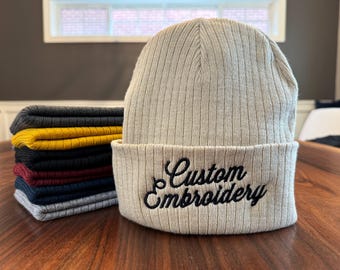 May include: A beige ribbed knit beanie with a black embroidered design that says "Custom Embroidery".