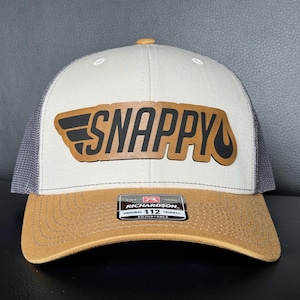 May include: A beige and brown trucker hat with a mesh back. The hat features the word "SNAPPY" in a brown leather patch with black accents. The brim is brown, and the hat has an adjustable strap.