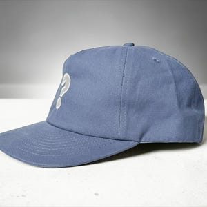 Custom embroidered question mark hat on blue cotton 5 panel snapback cap featuring structured crown and pre curved visor. Clean white embroidery symbol on front panel suitable for custom text, logo placement and daily wear. Adjustable snapback closure provides flexible fit with breathable cotton twill fabric. Product mockup shows front view on neutral background highlighting stitching detail, panel structure and consistent composition for all listing images.