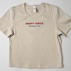 Custom embroidered baby tee for women in soft ribbed cotton, featuring personalized text on a fitted cropped American Apparel shirt. This trendy y2k baby tee showcases minimal embroidery design, perfect for aesthetic outfits, custom gifts, and everyday wear. Styled in a clean neutral background to highlight the flattering stretch fit, short sleeves, and modern cropped silhouette, ideal for fashion brands, casual styling, and personalized clothing collections.