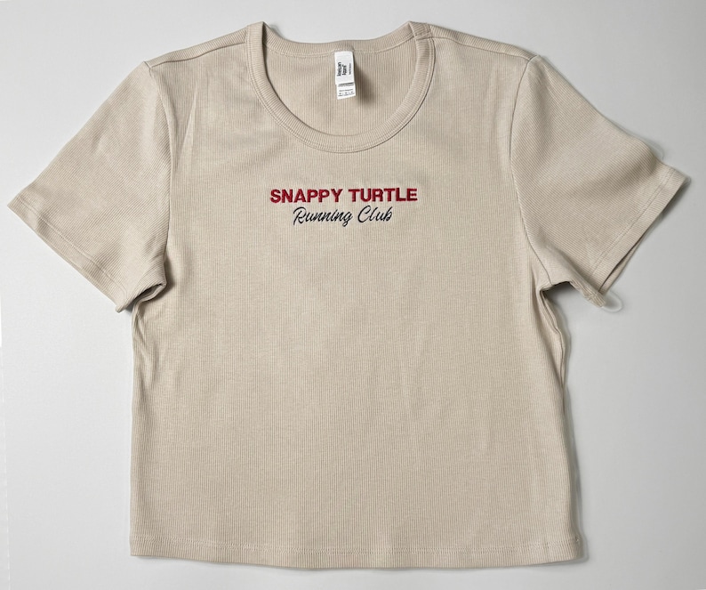 Custom embroidered baby tee for women in soft ribbed cotton, featuring personalized text on a fitted cropped American Apparel shirt. This trendy y2k baby tee showcases minimal embroidery design, perfect for aesthetic outfits, custom gifts, and everyday wear. Styled in a clean neutral background to highlight the flattering stretch fit, short sleeves, and modern cropped silhouette, ideal for fashion brands, casual styling, and personalized clothing collections.