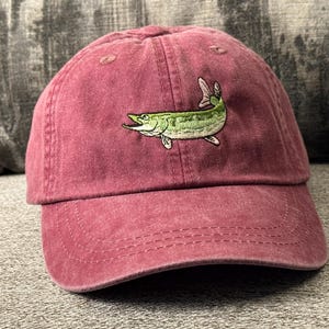 May include: A maroon baseball cap featuring an embroidered green and white fish design. The cap has a curved brim and a classic six-panel construction. The hat is made of a soft, textured fabric.