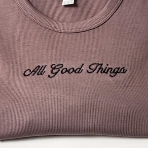 Custom embroidered baby tee for women in soft ribbed cotton, featuring personalized text on a fitted cropped American Apparel shirt. This trendy y2k baby tee showcases minimal embroidery design, perfect for aesthetic outfits, custom gifts, and everyday wear. Styled in a clean neutral background to highlight the flattering stretch fit, short sleeves, and modern cropped silhouette, ideal for fashion brands, casual styling, and personalized clothing collections.