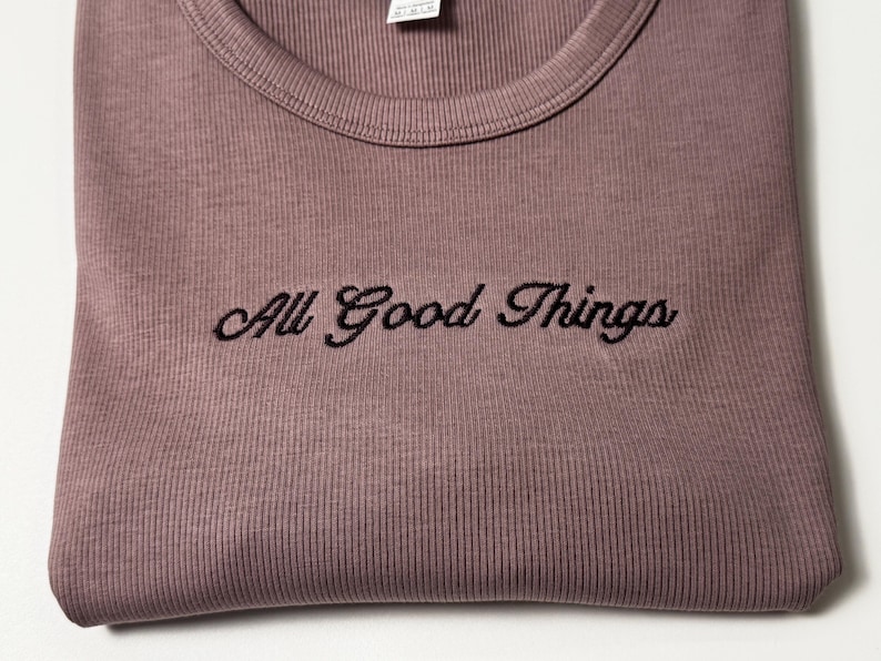 Custom embroidered baby tee for women in soft ribbed cotton, featuring personalized text on a fitted cropped American Apparel shirt. This trendy y2k baby tee showcases minimal embroidery design, perfect for aesthetic outfits, custom gifts, and everyday wear. Styled in a clean neutral background to highlight the flattering stretch fit, short sleeves, and modern cropped silhouette, ideal for fashion brands, casual styling, and personalized clothing collections.