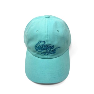 Custom embroidered script hat with personalized text on teal cotton twill dad cap featuring unstructured low profile shape and adjustable strap closure. Clean stitched lettering design on front panel suitable for branding, custom text display and everyday wear. Soft breathable cotton fabric with curved visor and detailed embroidery finish.