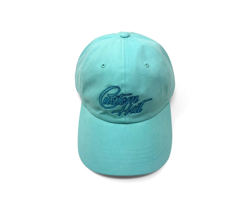 Custom embroidered script hat with personalized text on teal cotton twill dad cap featuring unstructured low profile shape and adjustable strap closure. Clean stitched lettering design on front panel suitable for branding, custom text display and everyday wear. Soft breathable cotton fabric with curved visor and detailed embroidery finish.