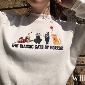 May include: White sweatshirt with a graphic of five cats in a row. The cats are wearing costumes and the text "The Classic Cats of Horror" is below them.