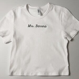 Custom embroidered baby tee for women in soft ribbed cotton, featuring personalized text on a fitted cropped American Apparel shirt. This trendy y2k baby tee showcases minimal embroidery design, perfect for aesthetic outfits, custom gifts, and everyday wear. Styled in a clean neutral background to highlight the flattering stretch fit, short sleeves, and modern cropped silhouette, ideal for fashion brands, casual styling, and personalized clothing collections.
