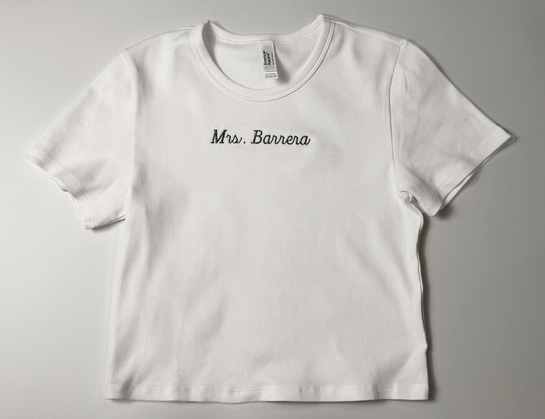 Custom embroidered baby tee for women in soft ribbed cotton, featuring personalized text on a fitted cropped American Apparel shirt. This trendy y2k baby tee showcases minimal embroidery design, perfect for aesthetic outfits, custom gifts, and everyday wear. Styled in a clean neutral background to highlight the flattering stretch fit, short sleeves, and modern cropped silhouette, ideal for fashion brands, casual styling, and personalized clothing collections.