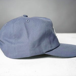 Custom embroidered question mark hat on blue cotton 5 panel snapback cap featuring structured crown and pre curved visor. Clean white embroidery symbol on front panel suitable for custom text, logo placement and daily wear. Adjustable snapback closure provides flexible fit with breathable cotton twill fabric. Product mockup shows front view on neutral background highlighting stitching detail, panel structure and consistent composition for all listing images.