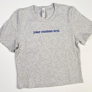 Custom embroidered baby tee for women in soft ribbed cotton, featuring personalized text on a fitted cropped American Apparel shirt. This trendy y2k baby tee showcases minimal embroidery design, perfect for aesthetic outfits, custom gifts, and everyday wear. Styled in a clean neutral background to highlight the flattering stretch fit, short sleeves, and modern cropped silhouette, ideal for fashion brands, casual styling, and personalized clothing collections.