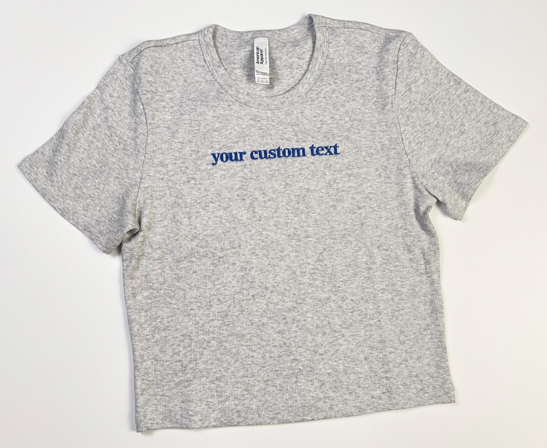 Custom embroidered baby tee for women in soft ribbed cotton, featuring personalized text on a fitted cropped American Apparel shirt. This trendy y2k baby tee showcases minimal embroidery design, perfect for aesthetic outfits, custom gifts, and everyday wear. Styled in a clean neutral background to highlight the flattering stretch fit, short sleeves, and modern cropped silhouette, ideal for fashion brands, casual styling, and personalized clothing collections.