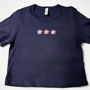 Custom embroidered baby tee for women in soft ribbed cotton, featuring personalized text on a fitted cropped American Apparel shirt. This trendy y2k baby tee showcases minimal embroidery design, perfect for aesthetic outfits, custom gifts, and everyday wear. Styled in a clean neutral background to highlight the flattering stretch fit, short sleeves, and modern cropped silhouette, ideal for fashion brands, casual styling, and personalized clothing collections.