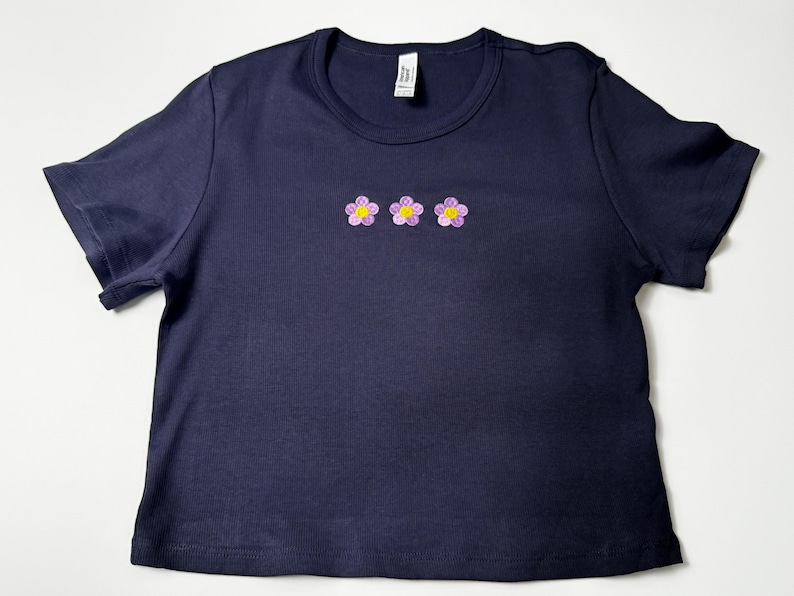 Custom embroidered baby tee for women in soft ribbed cotton, featuring personalized text on a fitted cropped American Apparel shirt. This trendy y2k baby tee showcases minimal embroidery design, perfect for aesthetic outfits, custom gifts, and everyday wear. Styled in a clean neutral background to highlight the flattering stretch fit, short sleeves, and modern cropped silhouette, ideal for fashion brands, casual styling, and personalized clothing collections.