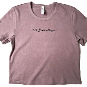 Custom embroidered baby tee for women in soft ribbed cotton, featuring personalized text on a fitted cropped American Apparel shirt. This trendy y2k baby tee showcases minimal embroidery design, perfect for aesthetic outfits, custom gifts, and everyday wear. Styled in a clean neutral background to highlight the flattering stretch fit, short sleeves, and modern cropped silhouette, ideal for fashion brands, casual styling, and personalized clothing collections.