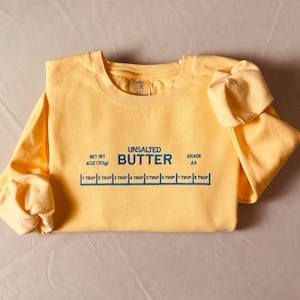May include: A yellow long-sleeved sweatshirt with the words "UNSALTED BUTTER" printed on the front, along with a butter measurement guide. The sweatshirt also has the text "NET WT 4 OZ (113g)" and "GRADE AA".
