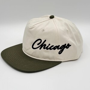 May include: A cream-colored baseball cap with a dark green brim and a matching button on top. The word "Chicago" is embroidered in black script across the front. This hat is a stylish accessory for casual wear.