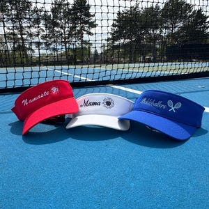 May include: Three visors in red, white, and blue are displayed on a blue surface. The red visor has "Namaste" embroidered on it. The white visor has "Mama" and a sunflower. The blue visor says "Pickeballers" with tennis rackets.