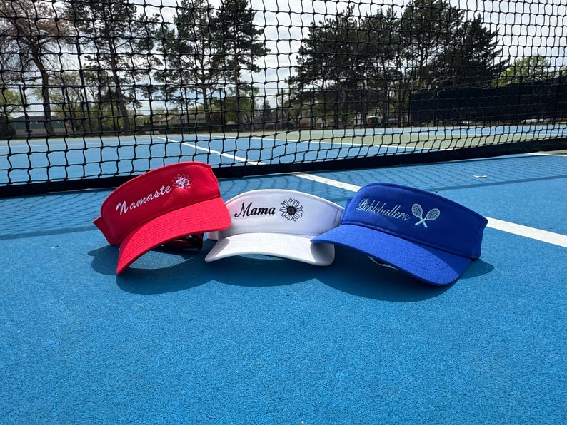 May include: Three visors in red, white, and blue are displayed on a blue surface. The red visor has "Namaste" embroidered on it. The white visor has "Mama" and a sunflower. The blue visor says "Pickeballers" with tennis rackets.