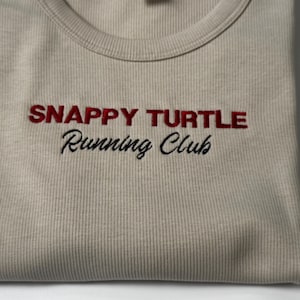 Custom embroidered baby tee for women in soft ribbed cotton, featuring personalized text on a fitted cropped American Apparel shirt. This trendy y2k baby tee showcases minimal embroidery design, perfect for aesthetic outfits, custom gifts, and everyday wear. Styled in a clean neutral background to highlight the flattering stretch fit, short sleeves, and modern cropped silhouette, ideal for fashion brands, casual styling, and personalized clothing collections.