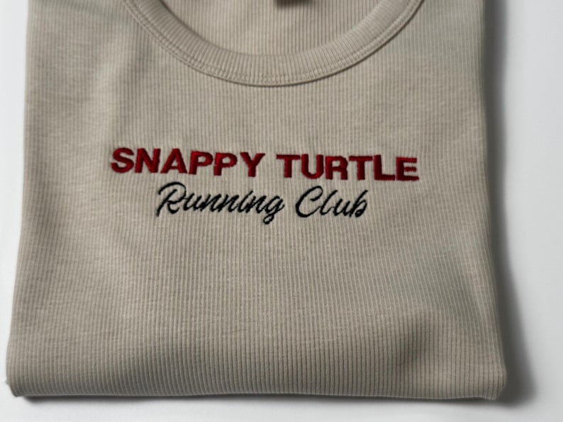 Custom embroidered baby tee for women in soft ribbed cotton, featuring personalized text on a fitted cropped American Apparel shirt. This trendy y2k baby tee showcases minimal embroidery design, perfect for aesthetic outfits, custom gifts, and everyday wear. Styled in a clean neutral background to highlight the flattering stretch fit, short sleeves, and modern cropped silhouette, ideal for fashion brands, casual styling, and personalized clothing collections.