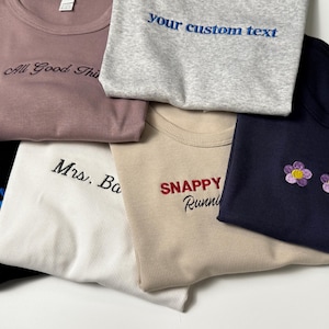 Custom embroidered baby tee for women in soft ribbed cotton, featuring personalized text on a fitted cropped American Apparel shirt. This trendy y2k baby tee showcases minimal embroidery design, perfect for aesthetic outfits, custom gifts, and everyday wear. Styled in a clean neutral background to highlight the flattering stretch fit, short sleeves, and modern cropped silhouette, ideal for fashion brands, casual styling, and personalized clothing collections.