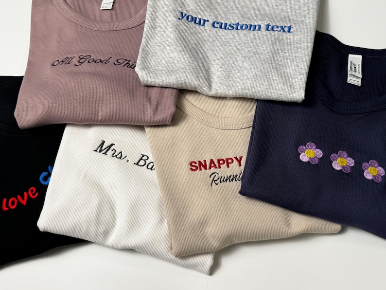Custom embroidered baby tee for women in soft ribbed cotton, featuring personalized text on a fitted cropped American Apparel shirt. This trendy y2k baby tee showcases minimal embroidery design, perfect for aesthetic outfits, custom gifts, and everyday wear. Styled in a clean neutral background to highlight the flattering stretch fit, short sleeves, and modern cropped silhouette, ideal for fashion brands, casual styling, and personalized clothing collections.
