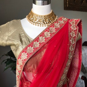 Op de afbeelding: Een rode en gouden sari met een gouden geborduurde rand. De sari is gedrapeerd over een mannequin en heeft een gouden ketting met parel accenten.