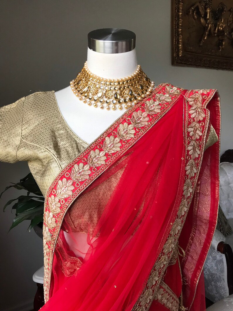 Op de afbeelding: Een rode en gouden sari met een gouden geborduurde rand. De sari is gedrapeerd over een mannequin en heeft een gouden ketting met parel accenten.