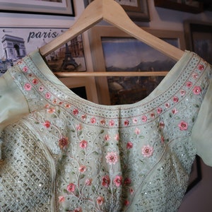 May include: A light green embroidered blouse with pink floral details and sequins. The blouse has a scoop neckline and is hanging on a wooden hanger.