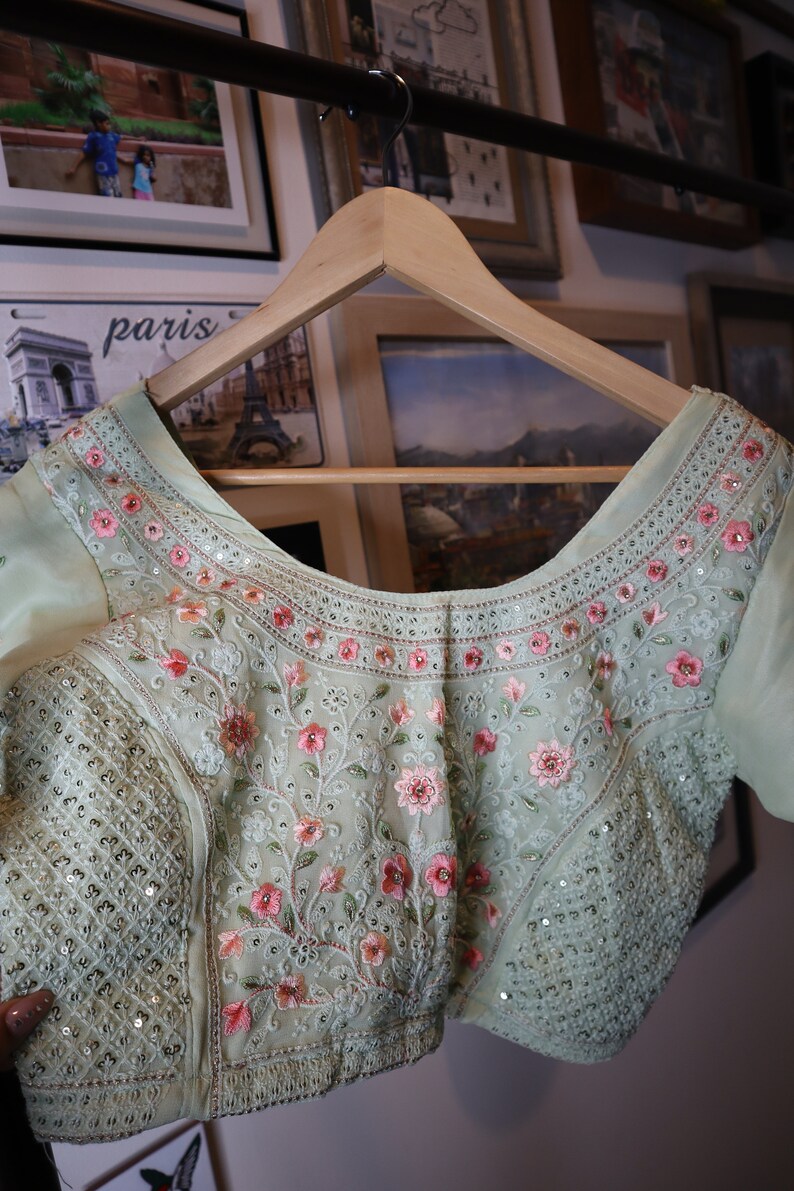 May include: A light green embroidered blouse with pink floral details and sequins. The blouse has a scoop neckline and is hanging on a wooden hanger.