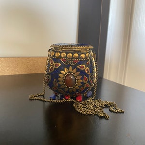 Puede incluir: Un peque&ntilde;o bolso de metal adornado en azul y dorado, con una correa de cadena. El bolso tiene una piedra roja y dise&ntilde;os florales intrincados.