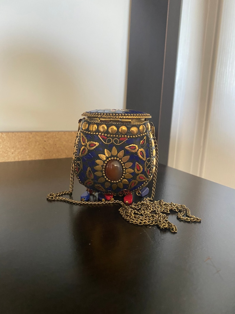 Puede incluir: Un peque&ntilde;o bolso de metal adornado en azul y dorado, con una correa de cadena. El bolso tiene una piedra roja y dise&ntilde;os florales intrincados.
