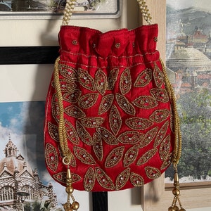 May include: A red drawstring bag with a gold beaded design and a pearl chain strap. The bag has a gold tassel on each side.