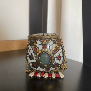 Puede incluir: Un peque&ntilde;o bolso redondo de mosaico blanco y negro con una cadena dorada y un borla rojo y blanco. El bolso tiene una piedra verde en el centro.