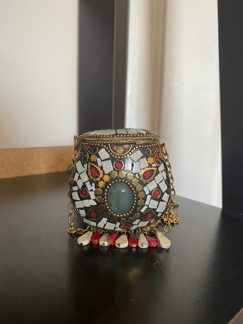 Puede incluir: Un peque&ntilde;o bolso redondo de mosaico blanco y negro con una cadena dorada y un borla rojo y blanco. El bolso tiene una piedra verde en el centro.