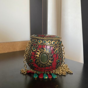 Puede incluir: Una peque&ntilde;a caja redonda roja y negra con detalles dorados y un borla turquesa y roja. La caja est&aacute; decorada con intrincados patrones y tiene una tapa con bisagras.